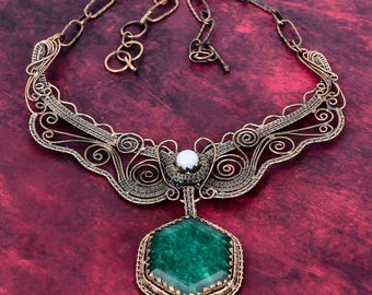 May include: A wire-wrapped necklace featuring a large hexagonal green stone pendant and a small white round bead. The necklace is crafted from dark copper-toned metal wire with intricate swirling patterns and a thick chain link closure.
