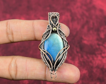 May include: A copper wire wrapped pendant featuring a blue labradorite gemstone. The pendant has a delicate, intricate design with a small gold bead accent.