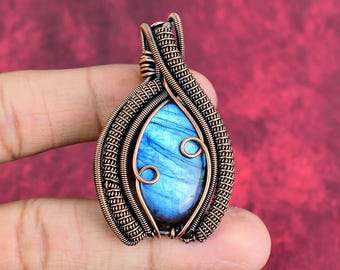 May include: A teardrop-shaped labradorite gemstone pendant encased in intricate, oxidized copper wire wrapping. The stone displays a vibrant blue iridescence, while the metalwork features detailed coils and spirals surrounding the central gem.