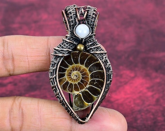 Puede incluir: Colgante artesanal con una concha de amonita fosilizada en un marco de alambre de cobre. El colgante presenta un patrón en espiral en tonos marrones y beige, con una pequeña cuenta blanca y una cuenta dorada en la parte superior.