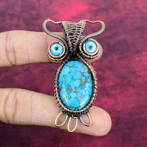 May include: A handcrafted copper wire pendant in the shape of an owl. The pendant features a turquoise oval stone at the centre, with blue eye-shaped accents. The copper wire is intricately woven around the stone and the eyes.