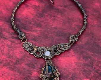 May include: A wire-wrapped necklace featuring a dark oxidized copper finish with intricate coiled metal patterns. The centerpiece includes a small, round, translucent white stone and a larger, teardrop-shaped dark blue stone with flecks.