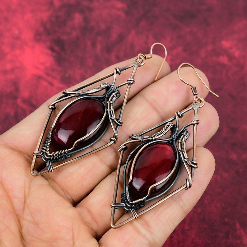 May include: A pair of dangling earrings with a geometric design. The earrings are made of copper wire and feature a red gemstone in the center of each earring.