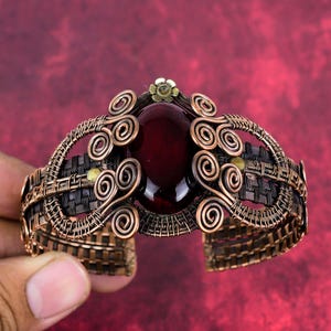 May include: A handcrafted copper cuff bracelet featuring a large, oval, deep red gemstone at its center. The bracelet has intricate wirework with spiral and woven patterns, and small gold-colored accents. The open design allows for easy wear.