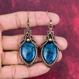 Puede incluir: Un par de pendientes envueltos en alambre de cobre con piedras preciosas de apatita azul. Los pendientes tienen un diseño delicado e intrincado y presentan un acento de cuentas doradas.