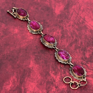 May include: A vintage bracelet featuring five oval, deep pink gemstones set in ornate, gold-toned metal frames. The bracelet has a clasp closure and is displayed against a textured, red background. The gemstones have a slightly rough texture.