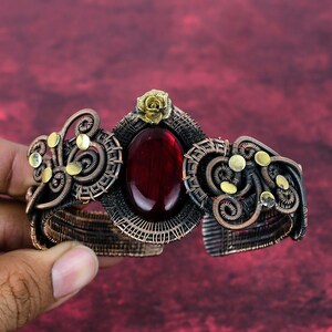 May include: A handcrafted copper wire cuff bracelet with a large oval red gemstone center. The bracelet features intricate wirework, a small gold-colored rose, and gold-colored accents. The overall design is ornate and detailed.