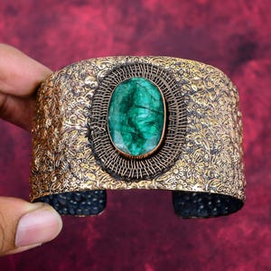 Op de afbeelding: Een brede, sierlijke manchetarmband met een grote, ovaalvormige groene edelsteen in het midden. De armband heeft een goudkleurig, bloemmotief en een donker, getextureerd interieur. De armband is ongeveer 5 cm breed.
