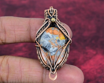 May include: A pendant featuring an oval-shaped stone with mottled orange, gray, and white patterns. The stone is encased in intricate, dark copper wire wrapping with a small gold-toned bead accent at the top and a circular loop at the base.