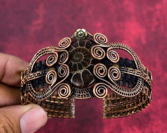 May include: A copper wire wrapped bracelet featuring a brown ammonite gemstone at its centre. The bracelet is adorned with copper wire spirals and has a woven copper band.