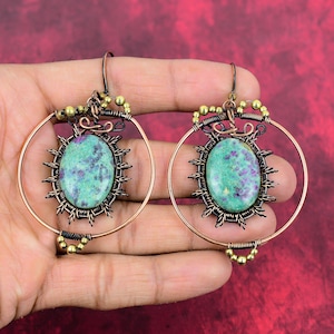 Peut inclure: Une paire de boucles d'oreilles créoles en cuivre avec une pierre précieuse verte et violette sertie dans un design en fil de fer. Les boucles d'oreilles pendent d'une main.