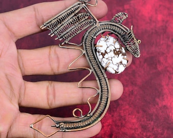 May include: A wire-wrapped pendant shaped like a dragon, crafted from dark copper-toned metal wire. The design features an oval-shaped white stone with brown dendritic inclusions set in the center, with intricate wire coils forming wings and a tail.