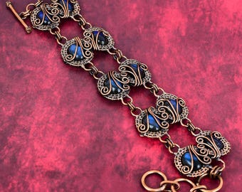 May include: A copper wire-wrapped bracelet featuring a series of oval-shaped dark blue iridescent stones. Each stone is encased in intricate copper wire coils and connected by copper jump rings, finished with a toggle clasp closure.