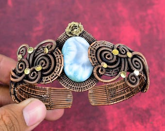 May include: A copper wire-wrapped cuff bracelet featuring a central oval light blue stone. The metalwork includes intricate swirls, small brass-colored circular accents, and a small brass-colored rose embellishment at the top center.