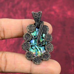 May include: Abalone shell pendant with a copper wire wrap. The iridescent shell is framed by a series of copper wire swirls. The pendant has a rectangular shape and is suspended from a copper wire bail.