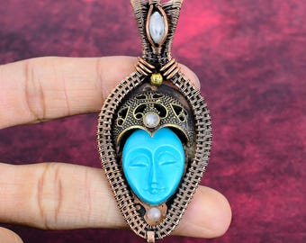 May include: A wire-wrapped copper pendant featuring a central carved turquoise-colored face mask, topped with a decorative filigree cap, and accented with three small white cabochon stones and a single small brass bead.