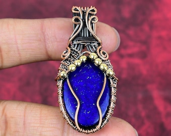 May include: A wire-wrapped pendant featuring an oval-shaped, deep blue lapis lazuli stone. The setting is crafted from dark oxidized copper wire with decorative copper coils and a row of small, polished gold-toned metal beads along the top.