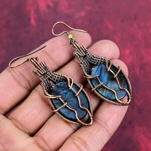 May include: A pair of copper wire wrapped earrings with oval shaped labradorite gemstones. The earrings have a dangling design and are a dark blue color.