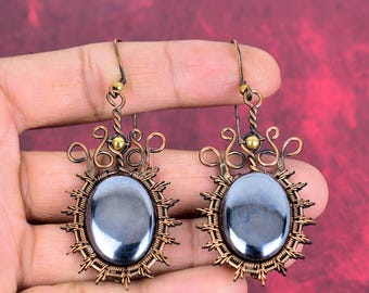 May include: A pair of dangle earrings featuring oval-shaped, polished dark gray hematite stones. The stones are encased in intricate, hand-wrapped copper wire frames with decorative swirls and small gold-toned metal beads at the top hooks.