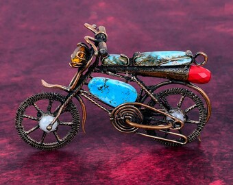 May include: A decorative motorcycle figurine crafted from copper-toned wire, featuring a bright blue oval stone on the frame, a long blue stone seat, a small amber headlight, a red tail light, and white circular accents in the center of each wheel.