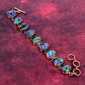 May include: A gold-toned bracelet featuring oval-shaped abalone shell links with iridescent blue and green hues. The bracelet has a toggle clasp and a series of connecting rings for closure. The jewelry is displayed on a red surface.