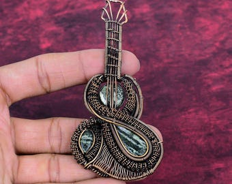 May include: A handcrafted copper wire guitar pendant with green stone accents. The pendant features intricate wire wrapping, creating a detailed miniature guitar design. The guitar pendant is held in a hand against a burgundy background.