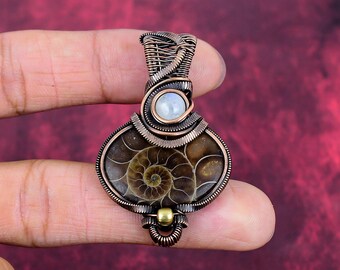Puede incluir: Colgante artesanal con un amonita fosilizada, encapsulada en alambre de cobre. El colgante incluye una pequeña piedra redonda de color claro y una cuenta dorada. El fósil de amonita muestra un patrón espiralado detallado.