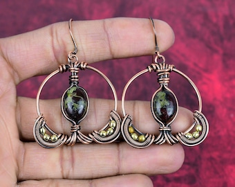 May include: A pair of copper wire earrings with a green and black stone in the center of each earring. The earrings are in the shape of a circle with a smaller circle inside. There are small gold beads around the edges of the earrings.
