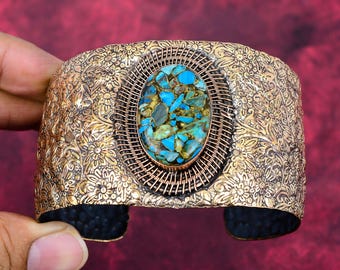 May include: A wide cuff bracelet featuring a textured, floral-patterned metallic surface in a bronze tone. The center displays an oval inlay of crushed turquoise and gold-colored stone fragments, surrounded by a detailed wire-wrapped border.