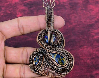 May include: A copper-colored wire-wrapped guitar pendant with blue and gold stone inlays. The pendant is held in a hand against a burgundy background. The guitar design features intricate wire detailing and a unique artistic style.