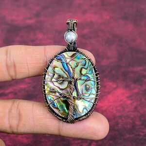 May include: Oval pendant with a tree of life design. It has a copper wire frame and a colourful abalone shell centre. A small, round, white stone is at the top. The pendant is held by a hand against a burgundy background.