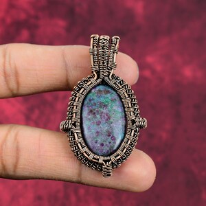 May include: A copper wire wrapped pendant featuring a green and purple oval gemstone. The pendant has a woven design and is held by a finger.
