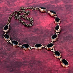 May include: A vintage-style necklace featuring oval black gemstones set in a gold-toned metal. The necklace has a chain and a pendant design, with a deep red background.