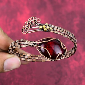 Puede incluir: Un brazalete de alambre de color cobre con una gema grande, ovalada y de color rojo oscuro en el centro. El brazalete presenta un intrincado trabajo de alambre y pequeñas cuentas doradas. El diseño incluye un elemento floral decorativo en la parte superior.
