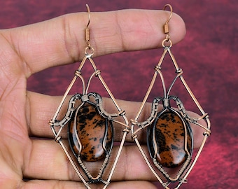 May include: A pair of dangle earrings featuring oval-shaped brown and black spotted stones encased in intricate copper wire-wrapped frames. The wirework forms a diamond-shaped structure around each stone, suspended from simple copper ear hooks.