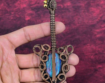 Puede incluir: Colgante de guitarra de alambre de cobre hecho a mano con incrustaciones de concha de abulón de colores. El colgante presenta un intrincado enrollado de alambre y tres cuentas doradas. El colgante en forma de guitarra se sostiene en una mano sobre un fondo burdeos.