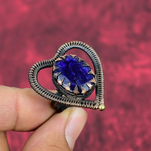 May include: A heart-shaped ring made of copper wire with a dark blue gemstone set in the center. The gemstone is surrounded by a copper frame with a decorative design.