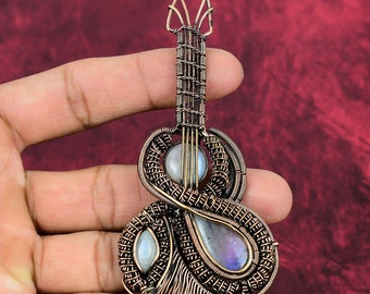 May include: A guitar-shaped pendant crafted from intricate copper wire wrapping. The design features three iridescent moonstone cabochons set within the wirework, including a round stone in the center and two teardrop-shaped stones at the base.