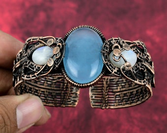 May include: A cuff bracelet crafted from dark oxidized copper wire woven into an intricate lattice pattern. The center features a large, oval, light blue cabochon stone, flanked by two smaller, round, translucent white moonstone cabochons.