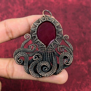 May include: A copper wire octopus pendant with a red gemstone cabochon in the center. The octopus has eight tentacles that are curled and detailed.