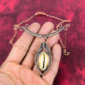 May include: A handcrafted copper necklace featuring a cowrie shell pendant. The pendant is encased in a wire-wrapped frame, suspended from a curved, woven wire design. The necklace has a chain with a lobster clasp.