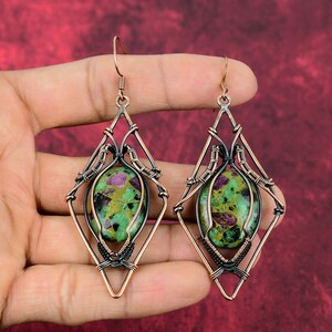 May include: A pair of handcrafted earrings featuring oval-shaped green and red gemstones set within a copper-colored wireframe. The earrings have a diamond-shaped design with intricate wire wrapping, and copper-colored ear wires. The background is a solid red.