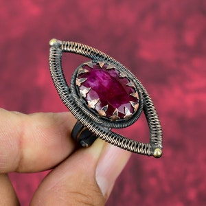 May include: A handcrafted ring featuring a large, oval-shaped, deep red gemstone. The gemstone is set in a copper-colored bezel with star-shaped accents. The ring has a unique, eye-shaped design with a wire-wrapped frame and a dark, oxidized finish.