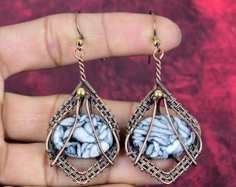 May include: A pair of dangle earrings featuring oval-shaped white stones with black veining, encased in intricate copper wire-wrapped frames. Each earring includes a small brass bead at the top and a hook-style closure for secure attachment.