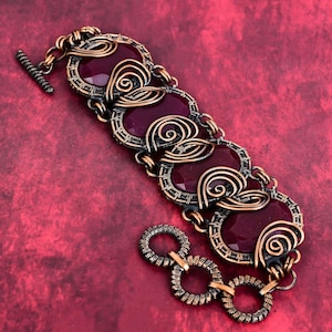 May include: A handcrafted bracelet featuring oval, deep red gemstones set in a copper-coloured wire frame. The bracelet has a toggle clasp and decorative wire swirls. The background is a textured, deep red surface.