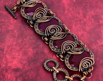 May include: A handcrafted bracelet featuring oval, deep red gemstones set in a copper-colored wire frame. The bracelet has a toggle clasp and decorative wire swirls. The background is a textured, deep red surface.