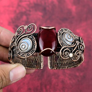 May include: Copper-toned cuff bracelet with a central, oval, deep red stone. The bracelet features intricate wirework with spiraling designs and two smaller, round, white stones. The open design allows for easy wear.