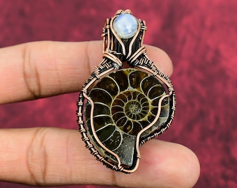 Puede incluir: Colgante artesanal con un amonita fosilizada envuelta en alambre de cobre. El patrón en espiral de la amonita es visible, con una pequeña piedra redonda e iridiscente en la parte superior. El colgante se muestra sobre un fondo rojo.