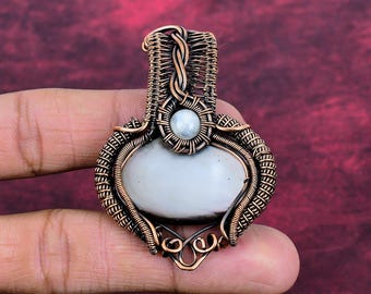 May include: A wire-wrapped pendant featuring a large, smooth, oval-shaped gray stone at the center, topped with a smaller, round, white iridescent bead. The entire piece is encased in intricate, coiled copper wire detailing.