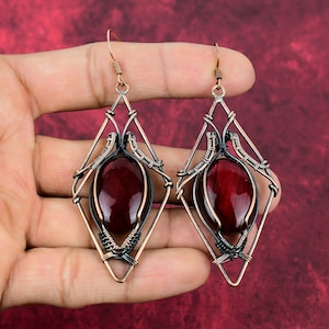 May include: A pair of dangle earrings with a geometric design. The earrings are made of copper wire and feature a red gemstone in the center. The wire is a mix of black and copper.